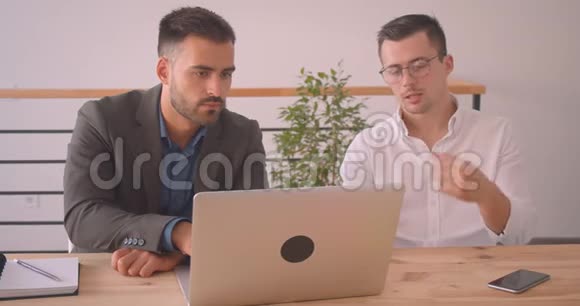 两位高加索商人在办公室室内一起讨论笔记本电脑项目的特写照片视频的预览图