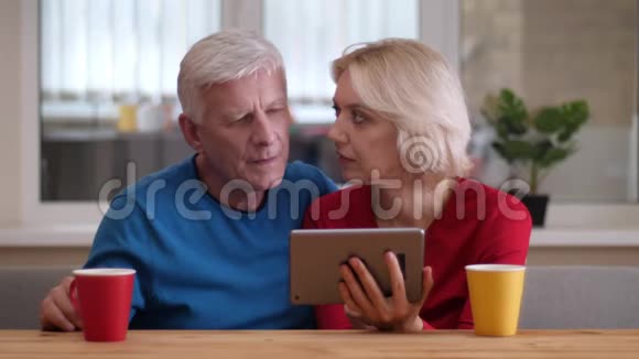 在一间舒适的公寓里一对年长的幸福夫妇在平板电脑上浏览特写镜头桌子上放着茶杯和茶视频的预览图