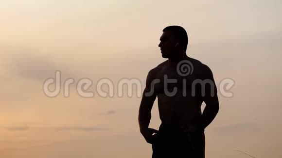 运动男子的剪影裸露的躯干摆姿势显示他的肌肉和二头肌在日落时靠在天空体育运动视频的预览图