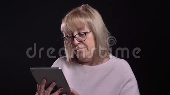 在镜头前的平板电脑上拍摄戴眼镜的白种人金发老奶奶的特写镜头视频的预览图