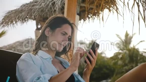 快乐轻松的旅行者女性使用智能手机电子商务应用程序微笑休息在海滩躺椅上异国情调的度假胜地视频的预览图