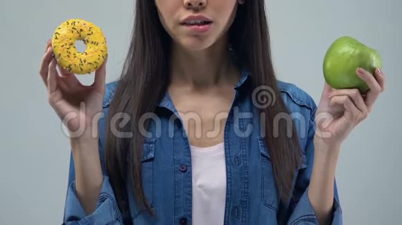 持有甜甜圈和苹果营养选择健康食品的困惑青年妇女视频的预览图