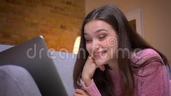 一位年轻漂亮的棕色白种人少女使用笔记本电脑在沙发上开心地笑着的特写镜头视频的预览图