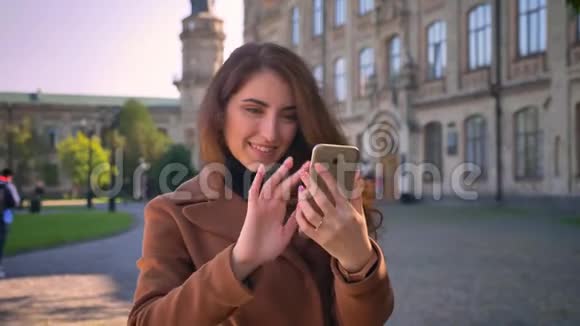 快乐的白种人黑发女人正在使用她的手机一只手拿着它一边站在城市里一边自拍视频的预览图