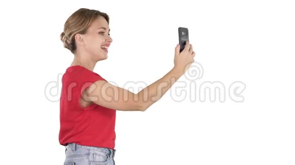 美丽的年轻女子走着拿着智能手机在白色背景上拍照和自拍视频的预览图