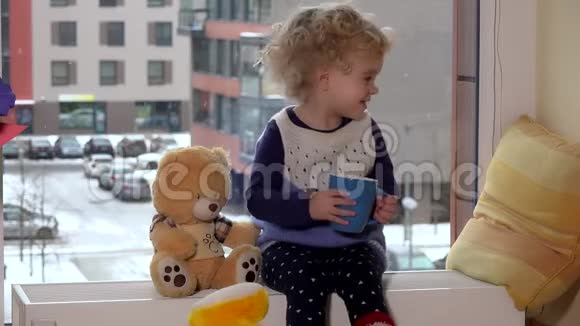 淘气的小女孩带着茶杯朋友泰迪熊坐在窗边视频的预览图