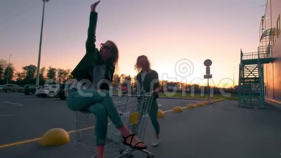 两个穿着街服的千禧女朋友在日落时在超市停车场玩得很开心坐着购物车视频的预览图