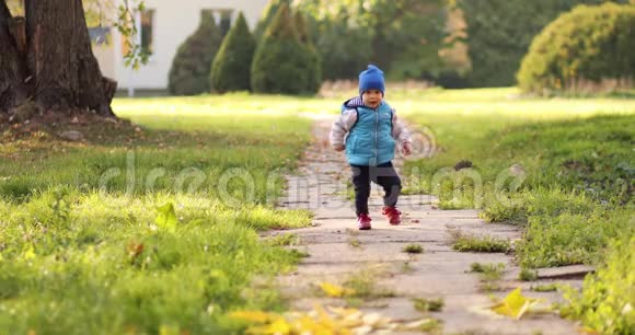 独自蹒跚学步的孩子戴着蓝色的帽子和背心在秋天的公园里滑稽地走开了视频的预览图