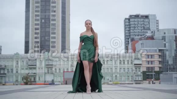 漂亮漂亮的女孩穿着一件迷人的晚绿色连衣裙在摩天大楼附近的空旷的城市广场上迷人地散步视频的预览图