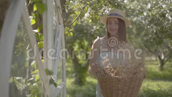 漂亮的年轻女子戴着草帽拿着柳条篮子拿着草药看着镜头在绿色的灯光下微笑视频的预览图