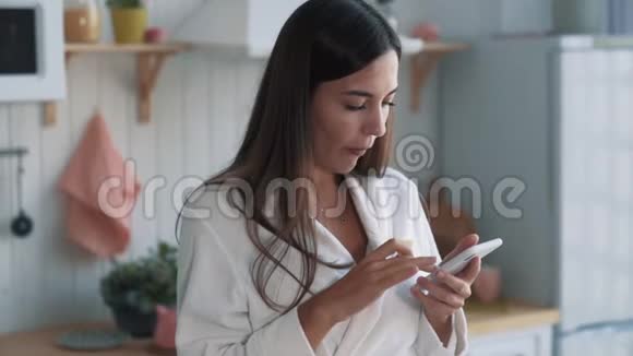 厨房里穿着浴袍的女孩的肖像吃苹果用电话慢动作视频的预览图
