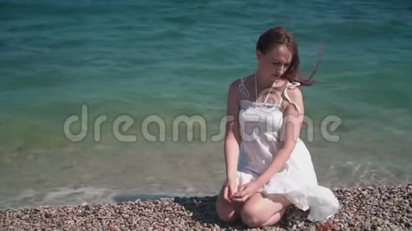 一个穿着白色海滩礼服的女孩坐在岩石海岸上一个漂亮的女孩坐在海滩上指着鹅卵石视频的预览图