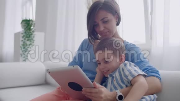 母子俩用平板电脑看着屏幕在舒适的房子里沙发上交谈视频的预览图