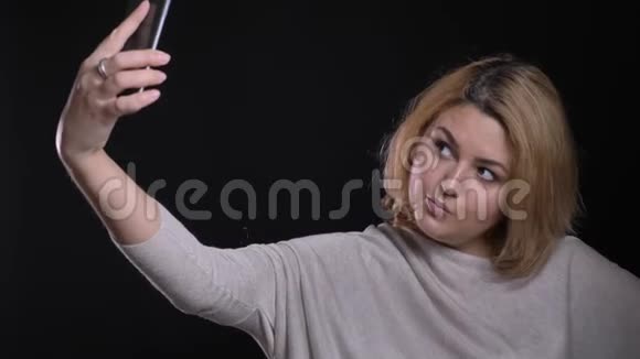 很高兴金发短发超重女商人在黑色背景下使用智能手机调情自拍视频的预览图