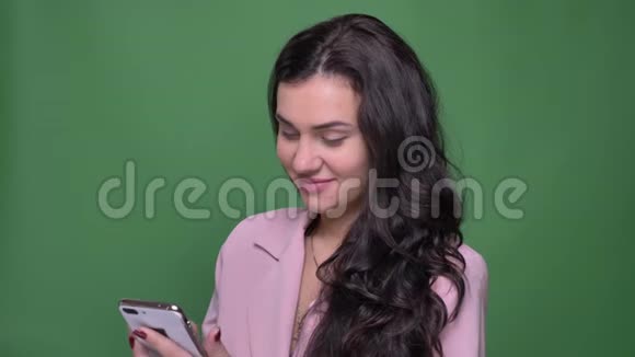 年轻的黑发女性vloger穿着粉红色夹克在绿色背景下微笑地看着智能手机的肖像视频的预览图
