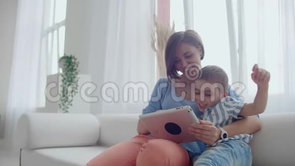 母子俩用平板电脑看着屏幕在舒适的房子里沙发上交谈视频的预览图