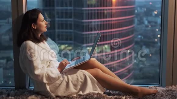 穿着浴袍的女孩在酒店的一个高楼层靠窗休息笔记本电脑后面的女博客视频的预览图
