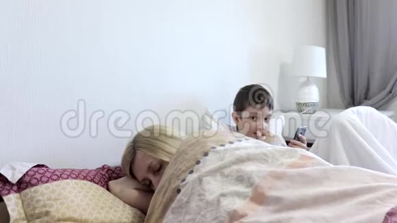 疲惫的母亲睡觉和她的太阳坐在床上玩手机游戏的小工具智能手机缺乏关注视频的预览图