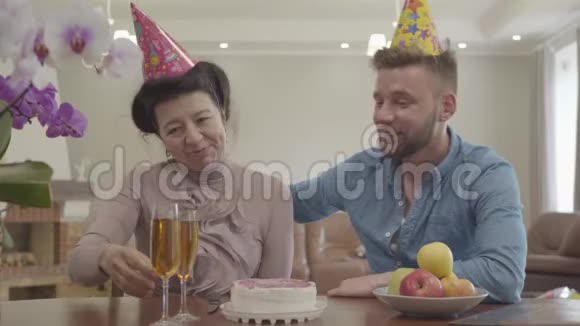 奶奶和成年孙子坐在桌边喝果汁头上戴着生日帽在桌子上视频的预览图