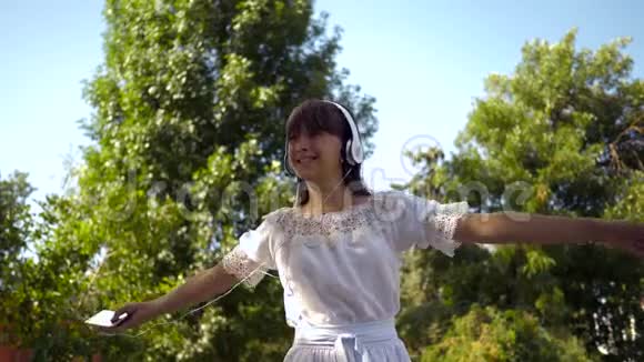 戴着耳机的快乐女孩在微笑手里拿着一部智能手机在夏天的公园里跳舞和旋转美丽美丽视频的预览图