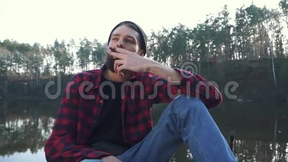 小胡子坐在河边的森林里抽着棕色的雪茄穿格子衬衫留着黑胡子的粗鲁男人视频的预览图