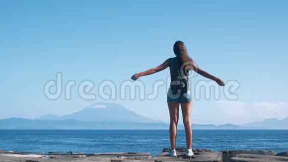 背着背包的女旅行者张开双臂欣赏大海火山的美景视频的预览图