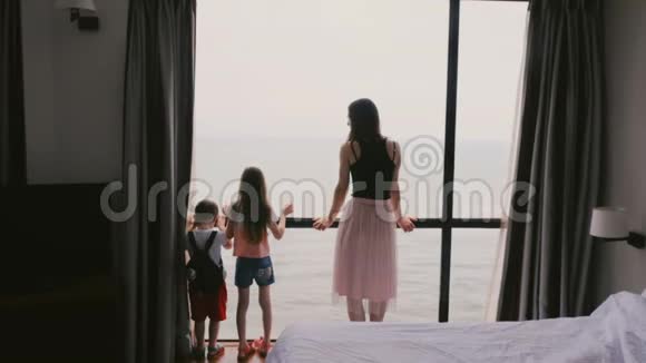 后景年轻妈妈和两个幸福的孩子站在一起在大酒店房间的窗户上盯着海景慢动作视频的预览图