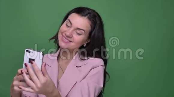 年轻的黑发vloger穿着粉红色夹克制作自拍照片并在绿色背景下的智能手机上观看视频的预览图