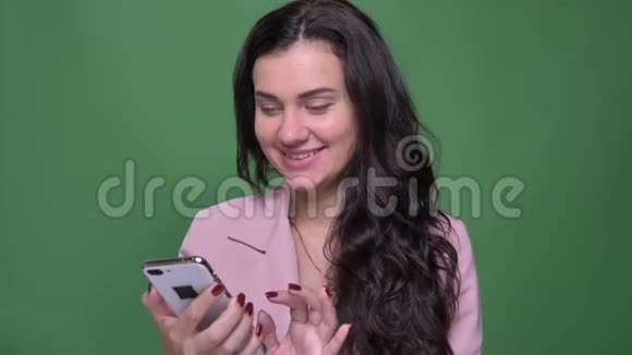 年轻的黑发女人穿着粉红色夹克在绿色背景下的智能手机上和朋友愉快地交谈视频的预览图