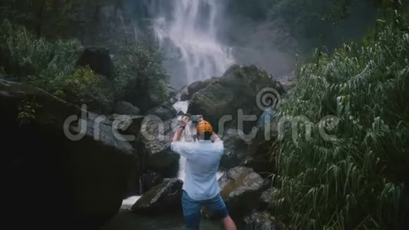 后景兴奋快乐的男性旅行博主在风景优美的斯里兰卡热带丛林瀑布拍摄智能手机视频视频的预览图