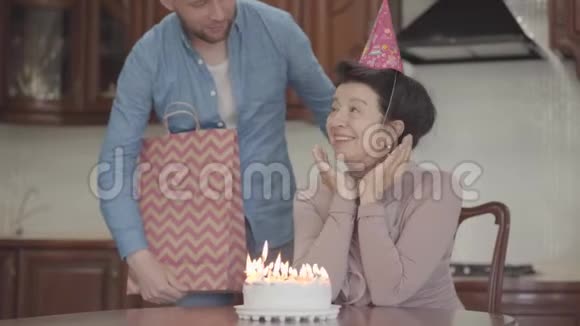 积极的情绪微笑的女士坐在桌子旁拿着小蛋糕和许多蜡烛成年孙子拥抱视频的预览图