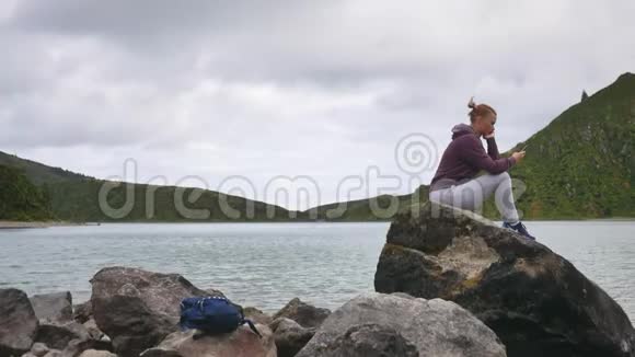 女孩坐在一个带有智能手机的山湖的背景上为旅行者使用应用程序视频的预览图