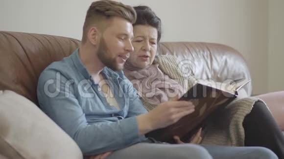 可爱的漂亮奶奶和成年孙子坐在棕色皮革沙发上看大照片视频的预览图
