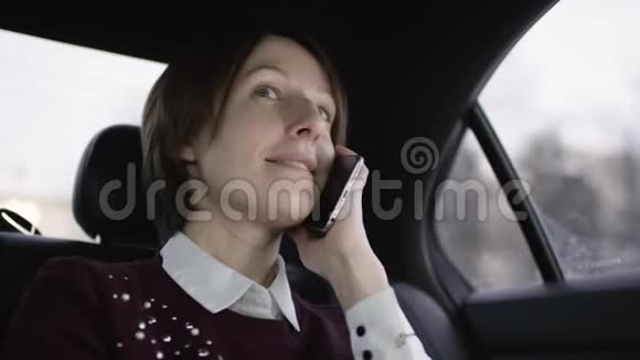 漂亮的女人棕色头发穿着白色衬衫和栗色毛衣坐在车里用电话说话视频的预览图