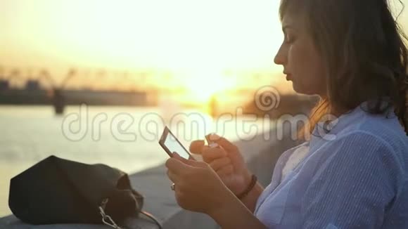 美丽的女人在河边夕阳的背景下看着一面小镜子用口红涂着嘴唇视频的预览图