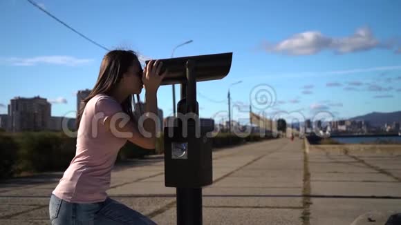 穿着粉红色衬衫和蓝色牛仔裤的旅行者年轻女子透过硬币固定望远镜观看视频的预览图