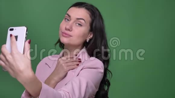 年轻的黑发女性vloger穿着粉色夹克用绿色智能手机制作自拍照片视频的预览图