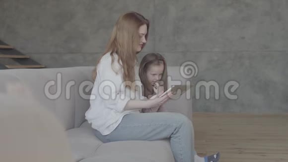 一位漂亮的年轻母亲和她可爱的小女儿正在用平板电脑微笑着坐在上面视频的预览图