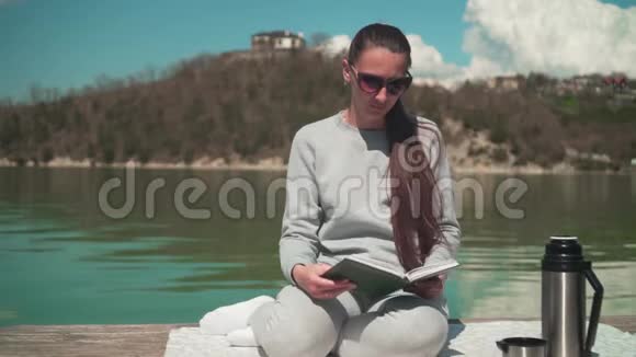 一个年轻的女人正在阳光下阅读一本书在春天的一天坐在一个湖的木墩上在大自然中放松视频的预览图