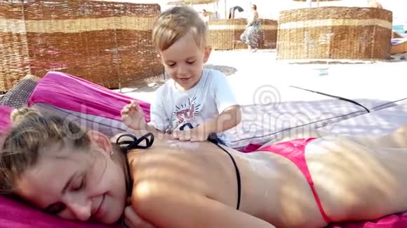 4k视频微笑的幼儿男孩涂防晒霜年轻母亲在海滨度假海滩放松视频的预览图