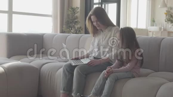 一位年轻的母亲和小女儿一头长发坐在客厅的沙发上用笔记本电脑打字视频的预览图