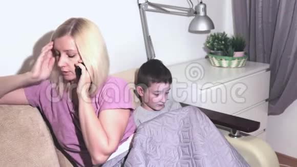 年轻的金发女人坐在沙发上通过电话说话一个男孩在后台玩智能手机视频的预览图
