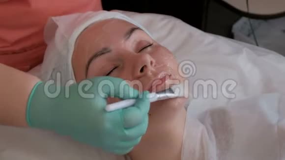 美容手术的漂亮女孩一位戴着蓝色手套的美容师在脸上涂上一层带有白色刷子的透明凝胶视频的预览图