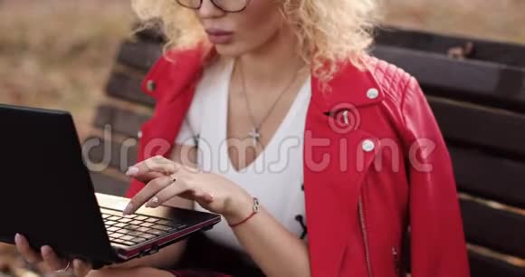成功的金发女人穿着明亮的休闲夹克在公园的笔记本电脑上打字视频的预览图