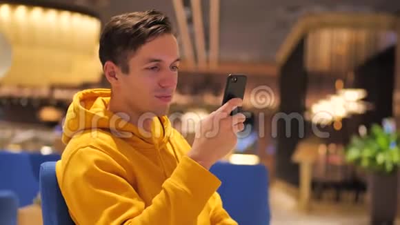 傍晚时分穿着黄色连帽衫的年轻人在舒适的咖啡馆里使用智能手机沟通商务旅行约会视频的预览图
