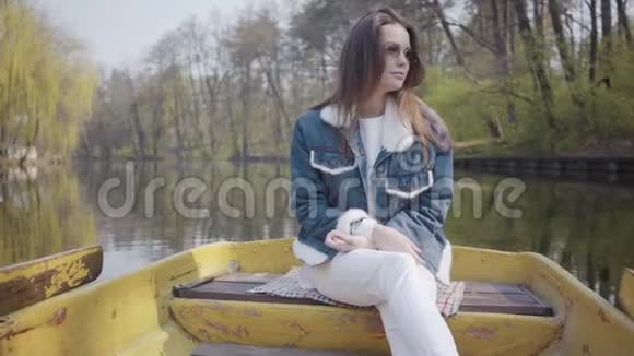 一个年轻漂亮的女人戴着太阳镜和一件牛仔夹克漂浮在湖或河上的船上的肖像美丽美丽视频的预览图