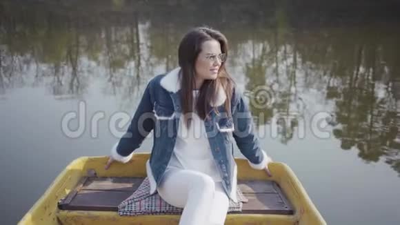 一个戴眼镜的漂亮女孩和一件牛仔夹克漂浮在湖或河上的船上的肖像美丽的黑发视频的预览图