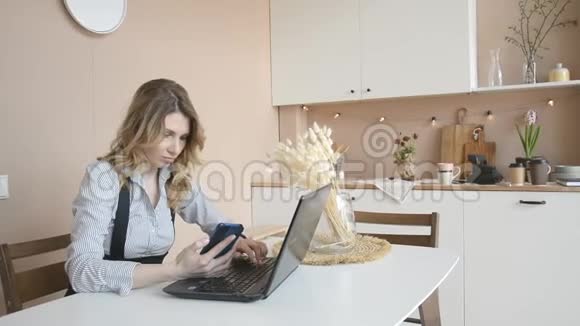 一个年轻漂亮的女人金发碧眼正坐在家里的厨房里用笔记本电脑工作视频的预览图