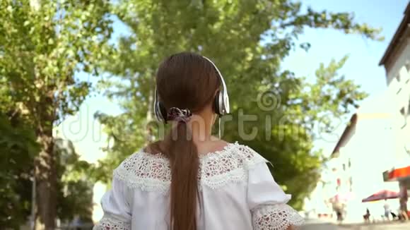 快乐的女孩戴着耳机听着音乐走在城市的街道上穿着白色长裙长发的女孩视频的预览图