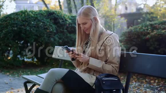微笑的女孩看着摸着电话坐在秋日的长凳上视频的预览图
